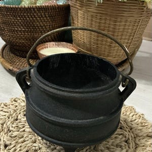 Op de afbeelding: Zwarte gietijzeren ketel met een handvat, staande op een geweven stro mat. De ketel heeft een ronde vorm en drie korte poten.