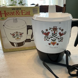 Puede incluir: Una olla eléctrica vintage Van Wyck Heat & Eat con cuerpo blanco, base y asa negras. La olla presenta un diseño floral con pájaros y el texto "Heat & Eat". También se ve la caja original.
