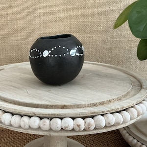 May include: A small, black, round vase with a wide opening. The vase is decorated with a white dotted pattern. It sits on a light-coloured, wooden, tiered stand with a beaded edge. The background is a textured, tan fabric.