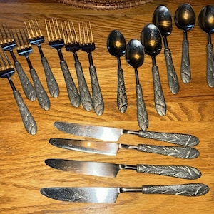 Puede incluir: Un conjunto de cubiertos antiguos, que incluye tenedores, cucharas y cuchillos, dispuestos sobre una superficie de madera. La cubertería presenta un detallado diseño de hojas en los mangos. Los cubiertos son de metal color plata.