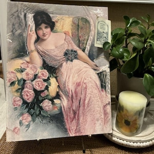 May include: A vintage illustration of a woman in a pink dress sitting in a chair with a bouquet of pink roses. The image is printed on a canvas and is displayed on a wooden easel.