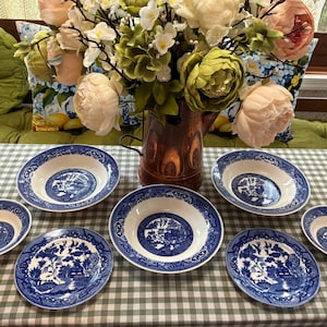 May include: A collection of white ceramic dinnerware featuring a blue willow pattern, including two large bowls, two smaller bowls, and two plates, arranged on a green and white checkered tablecloth with a copper pitcher and floral bouquet.
