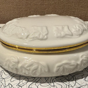 May include: A white ceramic jewellery box with a gold-coloured band around the middle. The box has a lid and base with a leaf design. The box is on a black and white patterned surface.