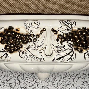Puede incluir: Una jardinera de cerámica blanca con un diseño de uvas y hojas en relieve en dorado y negro. La jardinera tiene una forma redondeada con un pie pequeño y un borde oscuro. Decorativa y vintage.