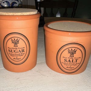 May include: Two terracotta kitchen canisters, one labeled "SUGAR" and the other "SALT." Both have cork lids and black and white labels with a floral design. The canisters are cylindrical and have a rustic, handmade appearance.