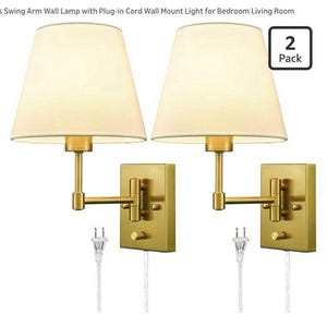 May include: Two gold-colored wall sconces with beige fabric shades. Each lamp has a swing arm design and a rectangular wall mount. The lamps are plugged in with white cords. The image includes a "2 Pack" label.