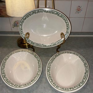 May include: Three off-white ceramic bowls with a green floral pattern along the rims. One bowl is displayed upright on a gold metal stand, while two identical bowls are positioned side-by-side in front of the stand on a flat surface.