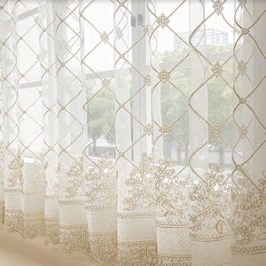 May include: Sheer white curtains featuring a gold embroidered lattice pattern across the upper section and a dense floral embroidery design along the bottom hem. The fabric is translucent, allowing light to pass through the intricate needlework.