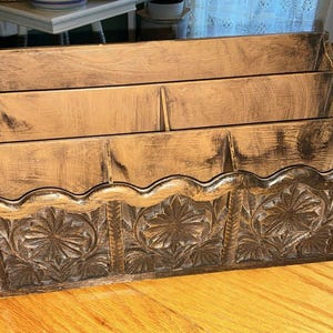 May include: A wooden desk organizer with three compartments and a decorative floral carved front. The organizer is stained in a warm brown color, with a scalloped edge along the bottom. The wood grain is visible.