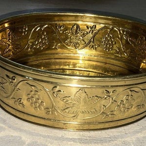 May include: A gold-colored, oval-shaped metal bowl with handles. The bowl features an embossed grape and leaf design around the rim. The bowl is approximately 10 inches long and 6 inches wide.