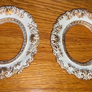 May include: Two ornate, oval picture frames with a white and gold finish. The frames have intricate floral and scrollwork details. The frames are displayed on a wooden surface.