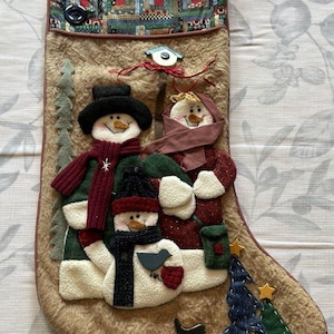 May include: A tan fuzzy Christmas stocking featuring three fabric snowmen figures, a green felt tree, and a patchwork cuff with two black buttons. The snowmen wear scarves, hats, and mittens, with additional small fabric trees and stars at the base.