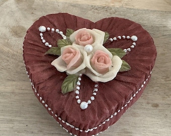 Op de afbeelding: Een hartvormige sieradendoos met een bordeauxrode fluwelen buitenkant en een witte parelrand. Het deksel heeft een delicaat bloemenmotief met drie roze en witte rozen.
