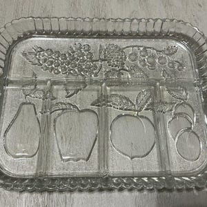 Puede incluir: Bandeja de servir de vidrio transparente con forma rectangular y diseño de frutas decorativo. La bandeja presenta imágenes en relieve de uvas, peras, manzanas, melocotones y cerezas. Tiene un borde festoneado y asas con detalles redondos.
