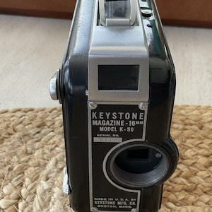 May include: Black Keystone Magazine-16mm Model K-50 camera with a silver top and a black leather handle. The camera has a lens and a label with text. The serial number is 2270. Made in the U.S.A.