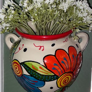 May include: A cream-colored ceramic wall planter featuring two side handles and a vibrant hand-painted floral design in orange, blue, green, and yellow. The rim is painted bright red, and the planter is filled with a bouquet of small white flowers.