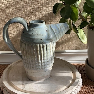 Puede incluir: Una jarra de cerámica hecha a mano con un esmalte azul claro y blanco. La jarra tiene un asa grande y un pico. El cuerpo de la jarra tiene un diseño texturizado y acanalado. Se encuentra sobre una bandeja redonda de madera.