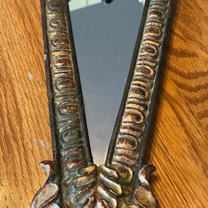 May include: An antique-style, V-shaped mirror with an ornate, bronze-colored frame. The frame features detailed scrollwork and a decorative base. The mirror's surface is reflective, and the overall design suggests a vintage aesthetic.