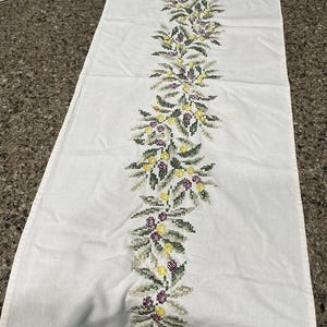May include: White table runner with a central embroidered floral design. The embroidery features green leaves, yellow berries, and purple accents. The runner is made of a soft, white fabric.
