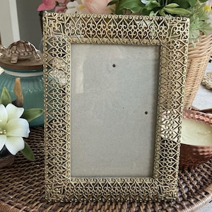 May include: Ornate gold-colored picture frame with a detailed filigree design. The rectangular frame has a neutral-colored backing and is displayed on a woven tray. Decorative elements include a turtle figurine and a white flower.
