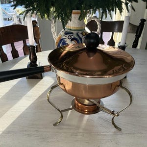 Può includere: Scaldavivande in rame con manico e coperchio neri. Il piatto ha una fascia bianca al centro e poggia su un supporto in ottone. Un pomello nero sovrasta il coperchio. Lo sfondo comprende candele e un vaso decorativo.