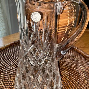 May include: A clear crystal pitcher with a diamond-cut pattern and a curved handle. The pitcher has a wide base and a flared top. A small, round sticker is visible on the front. The pitcher is sitting on a woven tray.