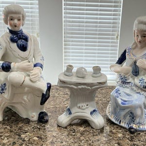 May include: A set of three porcelain figurines depicting a man and woman in period clothing, with a small table. The figures are white with blue accents and floral patterns. The table has a white base and a round top with small tea cups.