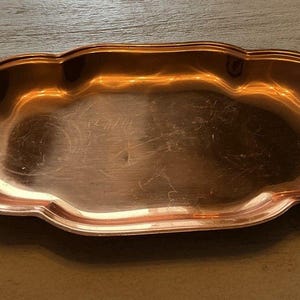 May include: A copper-colored, decorative tray with a scalloped edge. The tray has a smooth, reflective surface and a shallow, oval shape. The copper finish gives the tray a warm, antique appearance. This tray is suitable for serving or display.