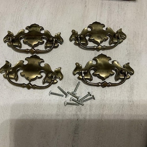 May include: Four antique brass drawer pulls with ornate designs, arranged on a light-colored surface. Each pull has a curved handle and decorative detailing. A collection of small screws is also present, suggesting a hardware set.