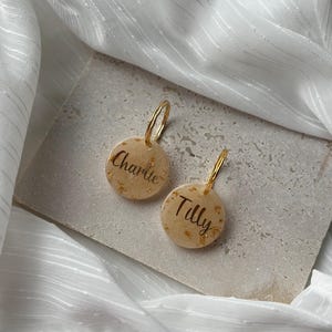 Könnte beinhalten: Zwei personalisierte goldene Creolen. Jeder Ohrring hat einen runden, beigefarbenen Anhänger mit Goldflocken und den Namen "Charlie" und "Tilly" in schwarzer Schrift. Die Ohrringe liegen auf einer steinernen Oberfläche.