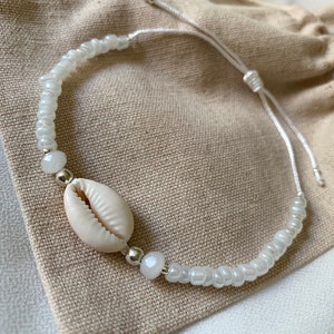 Könnte beinhalten: Weißes Perlenarmband mit einem weißen Muschel-Anhänger und silbernen Perlen. Das Armband liegt auf einem hellbraunen Stoffhintergrund.