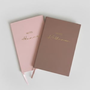 Könnte beinhalten: Zwei rechteckige Notizbücher mit einfarbigen Einbänden in Hellrosa und gedecktem Braun. Jedes Notizbuch trägt das Wort NOTES in Gold über einem goldenen, kursiven Namen, Hanna auf dem rosa und Katharina auf dem braunen Einband, mit Lesebändchen.