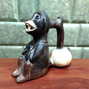 May include: A black ceramic monkey figurine with a white face and a round, white ceramic base. The monkey is sitting with its legs crossed and has a spout on its back.