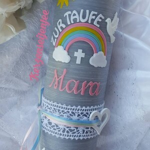 May include: A gray candle with a rainbow, a dove, a sun, a cross, and the name "Mara" in pink lettering. The date "4.10.2020" is written on the candle. The candle is decorated with white lace and ribbons.
