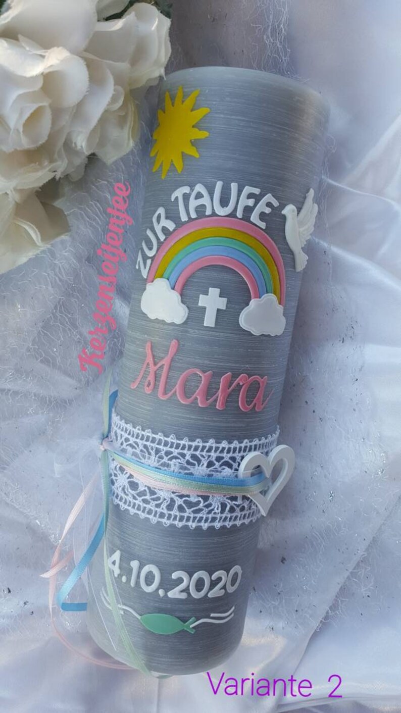 May include: A gray candle with a rainbow, a dove, a sun, a cross, and the name "Mara" in pink lettering. The date "4.10.2020" is written on the candle. The candle is decorated with white lace and ribbons.