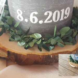 May include: A gray candle with the date 8.6.2019 written on it. The candle is sitting on a wooden slice with a green garland around the base.