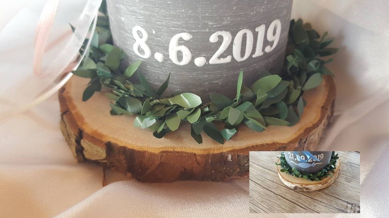 May include: A gray candle with the date 8.6.2019 written on it. The candle is sitting on a wooden slice with a green garland around the base.