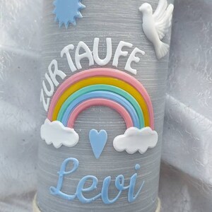 May include: A gray candle with a white dove, a blue star, a rainbow, and clouds. The text "Zur Taufe Levi" is written on the candle. The candle is decorated with a white rope and a wooden anchor.