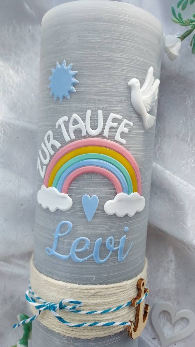 May include: A gray candle with a white dove, a blue star, a rainbow, and clouds. The text "Zur Taufe Levi" is written on the candle. The candle is decorated with a white rope and a wooden anchor.