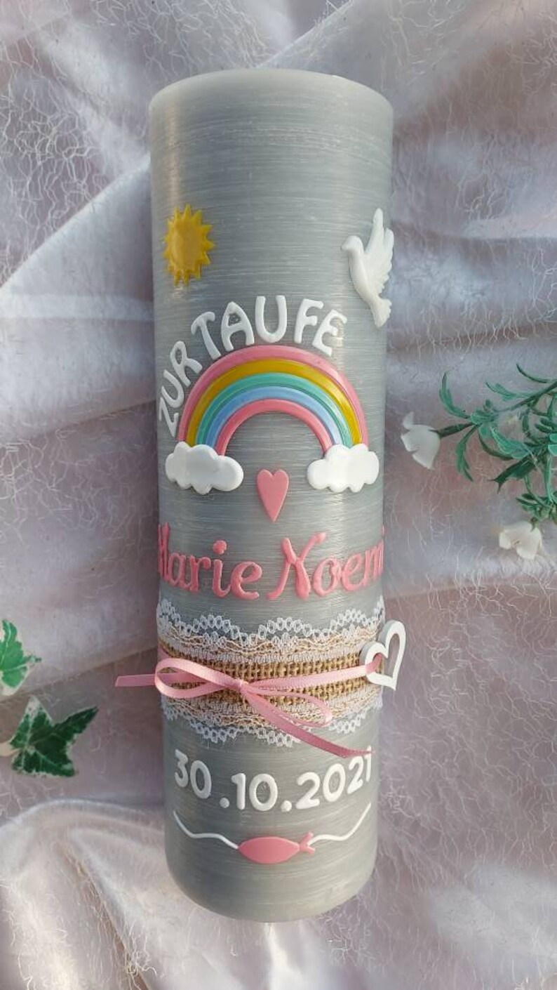May include: Gray baptism candle with a white dove, a rainbow, and a pink heart. The candle is decorated with a white lace ribbon and a pink ribbon. The text "Zur Taufe Marie Noem 30.10.2021" is written on the candle.