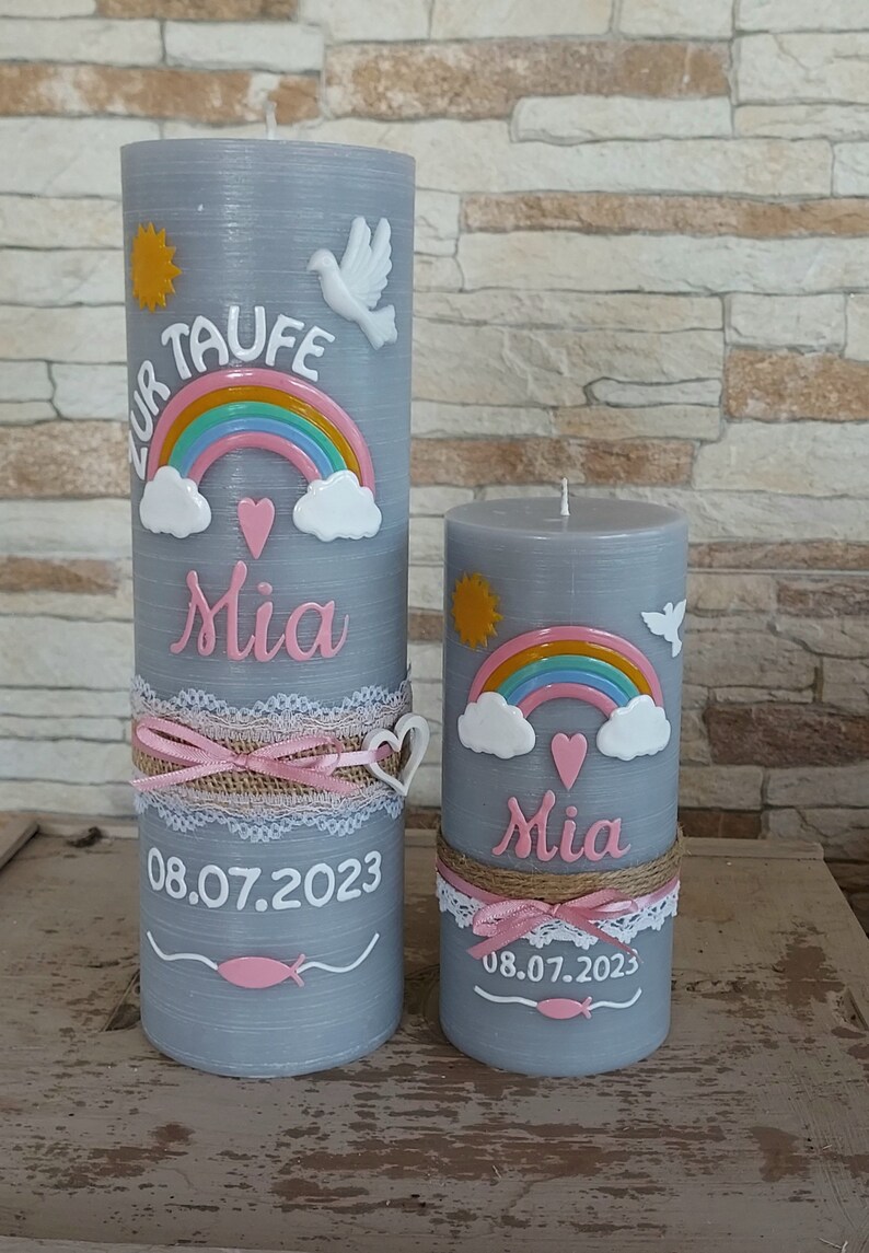 May include: Two gray pillar candles with a rainbow, clouds, and a heart design. The larger candle has the text "Zur Taufe Mia 08.07.2023" and the smaller candle has the text "Mia 08.07.2023".