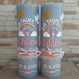 May include: Two gray baptism candles with a rainbow, clouds, a heart, a dove, and a sun design. The candles are decorated with a lace ribbon and a pink bow. The text "Zur Taufe" is written on each candle, along with the names "Chiara" and "Catalea" and the date "17.9.2023".