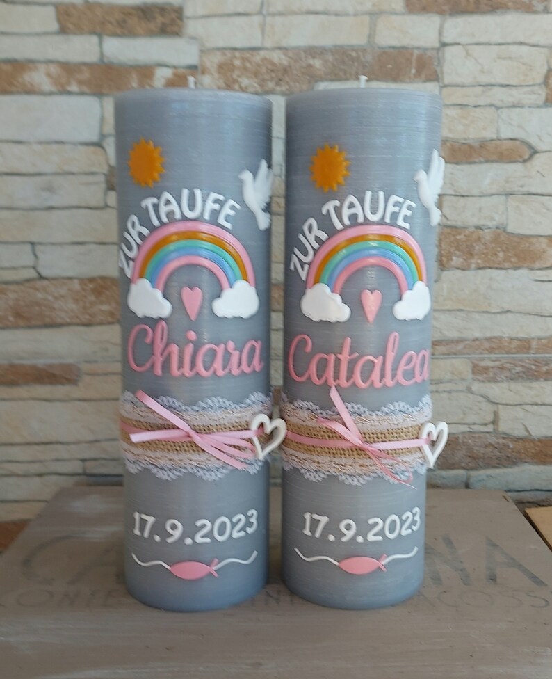 May include: Two gray baptism candles with a rainbow, clouds, a heart, a dove, and a sun design. The candles are decorated with a lace ribbon and a pink bow. The text "Zur Taufe" is written on each candle, along with the names "Chiara" and "Catalea" and the date "17.9.2023".