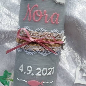 May include: A gray candle with a pink and white rainbow, clouds, and a heart. The name "Nora" is written in pink lettering. The date "4.9.2021" is written in white lettering. A pink ribbon and lace are wrapped around the candle.