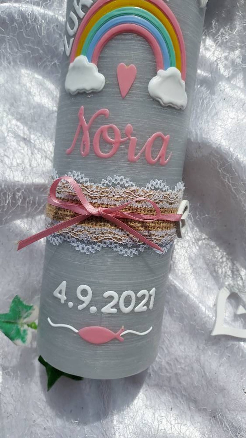 May include: A gray candle with a pink and white rainbow, clouds, and a heart. The name "Nora" is written in pink lettering. The date "4.9.2021" is written in white lettering. A pink ribbon and lace are wrapped around the candle.