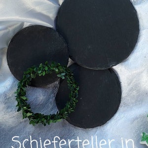 May include: Three round black slate serving plates of different sizes on a white fabric background. The text "Schieferteller in verschiedenen Gr&ouml;&szlig;en" is written in white on the fabric.