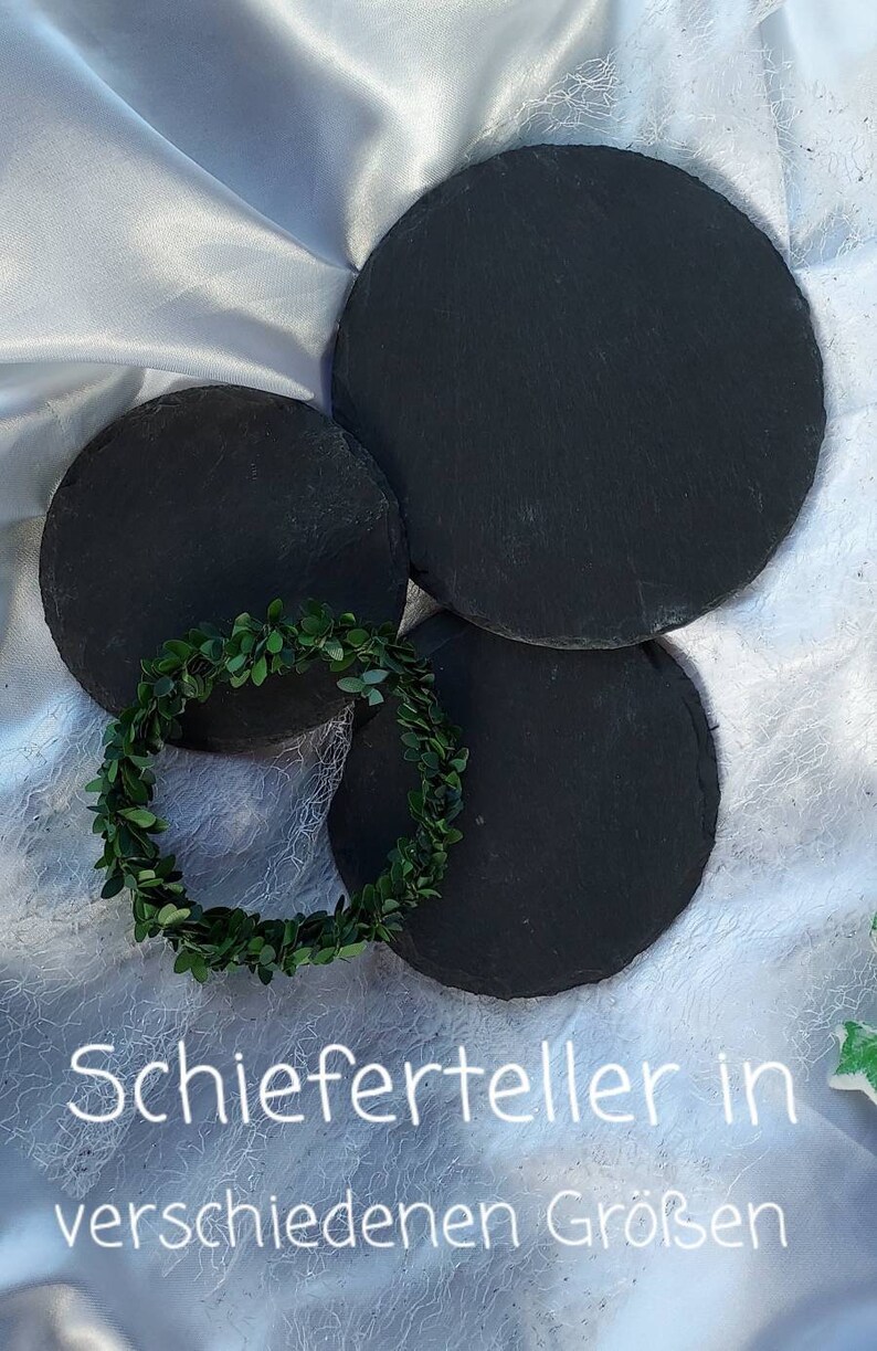 May include: Three round black slate serving plates of different sizes on a white fabric background. The text "Schieferteller in verschiedenen Gr&ouml;&szlig;en" is written in white on the fabric.