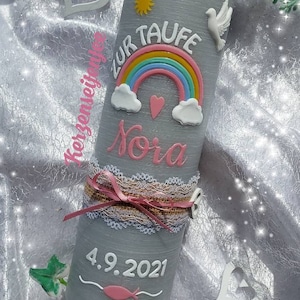 May include: Gray baptism candle with a rainbow, clouds, a heart, a dove, and a sun. The candle is decorated with a white lace ribbon and a pink bow. The text "Zur Taufe Nora 4.9.2021" is written on the candle.
