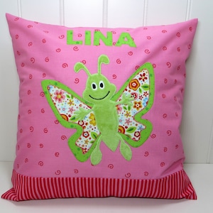 Könnte beinhalten: Rosa quadratisches Kissen mit einem grünen Schmetterling-Applikation. Der Schmetterling hat Flügel mit Blumenmuster und ein lächelndes Gesicht. Das Wort "LINA" ist in grünen Buchstaben gestickt. Roter und weißer Streifen am Rand.