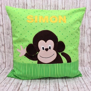 Könnte beinhalten: Ein quadratisches grünes Kissen mit einem lächelnden Affen-Applikation. Der Affe ist braun mit einem hellrosa Gesicht und winkt. Der Name "SIMON" ist gelb über dem Affen gestickt. Das Kissen hat einen grün gestreiften Rand.